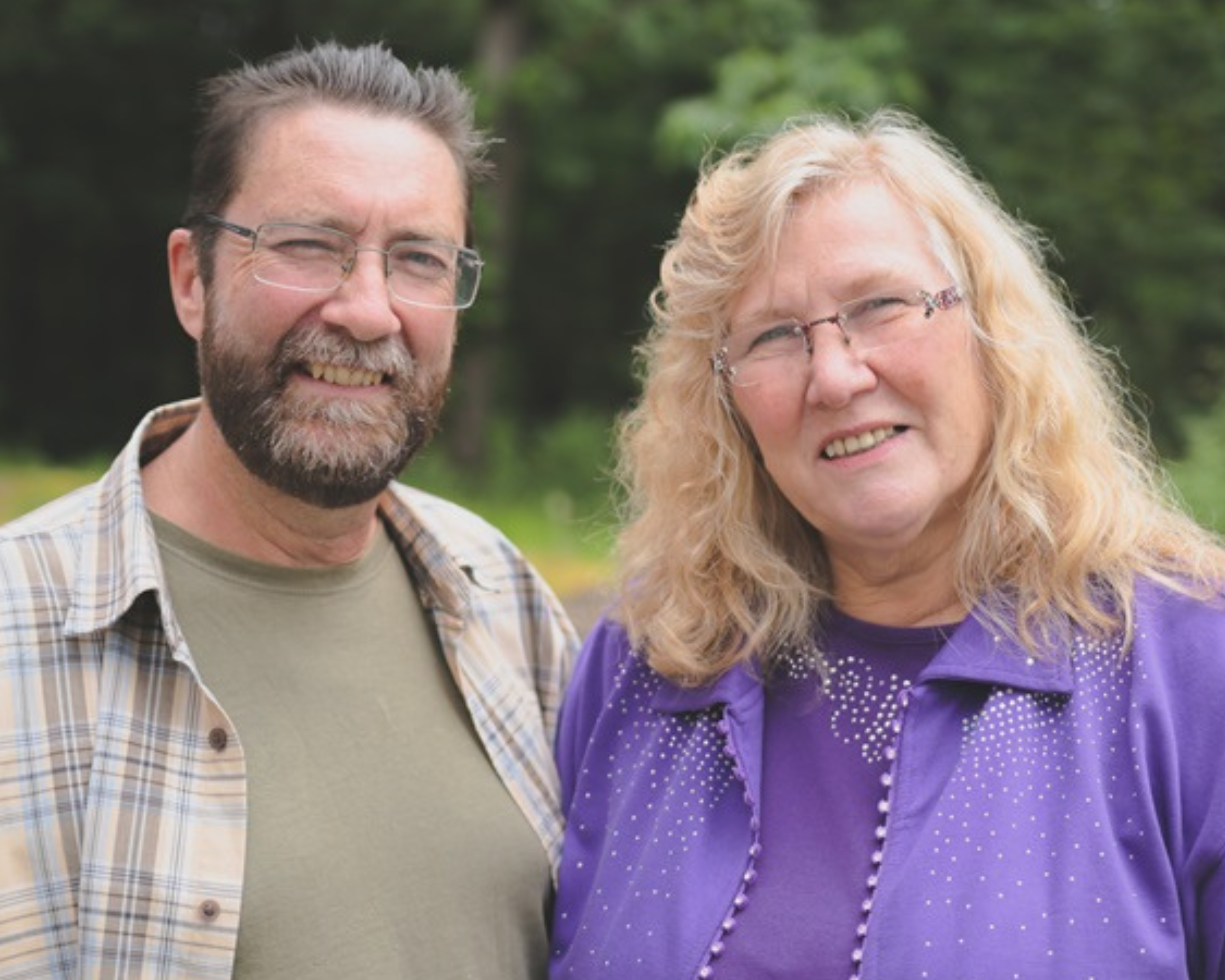 A middle-aged couple stands together outdoors, smiling at the camera. The man on the left is wearing glasses, a beard, and a plaid shirt over a green t-shirt. The woman on the right has long, wavy blonde hair and is wearing glasses and a purple jacket adorned with small decorative beads. The background is lush and green, suggesting a natural, wooded setting, which adds to the peaceful and relaxed atmosphere of the photo.