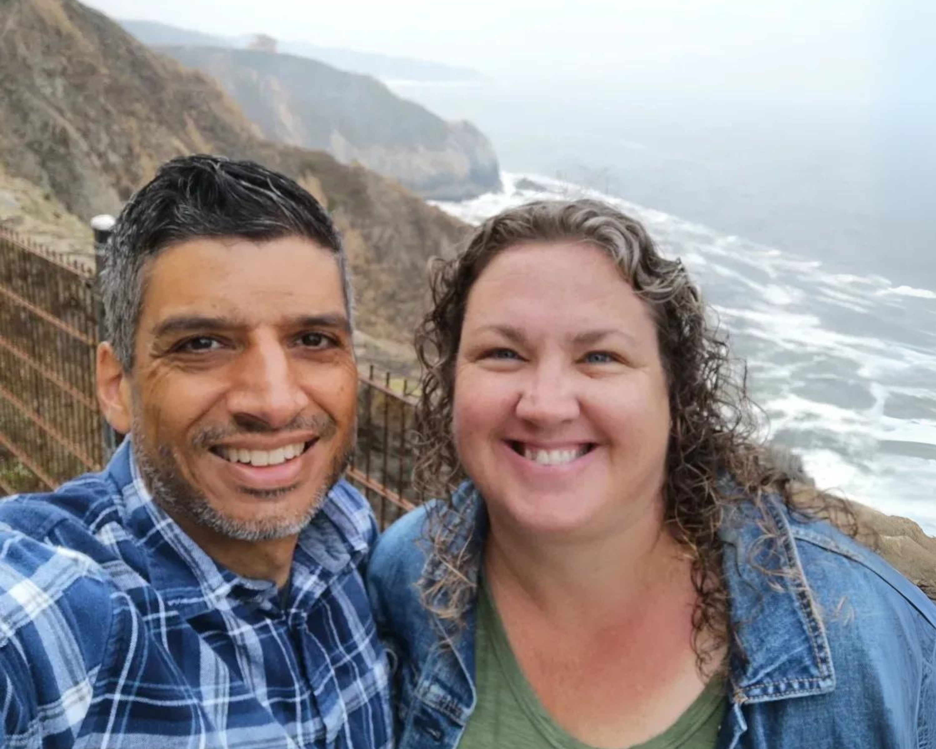 A smiling couple stands together on a cliff overlooking a foggy coastline. The man on the left is wearing a blue plaid shirt, and the woman on the right is wearing a green top with a denim jacket. The ocean waves crash against the rocky shore below, and the fog adds a misty, serene atmosphere to the scene.
