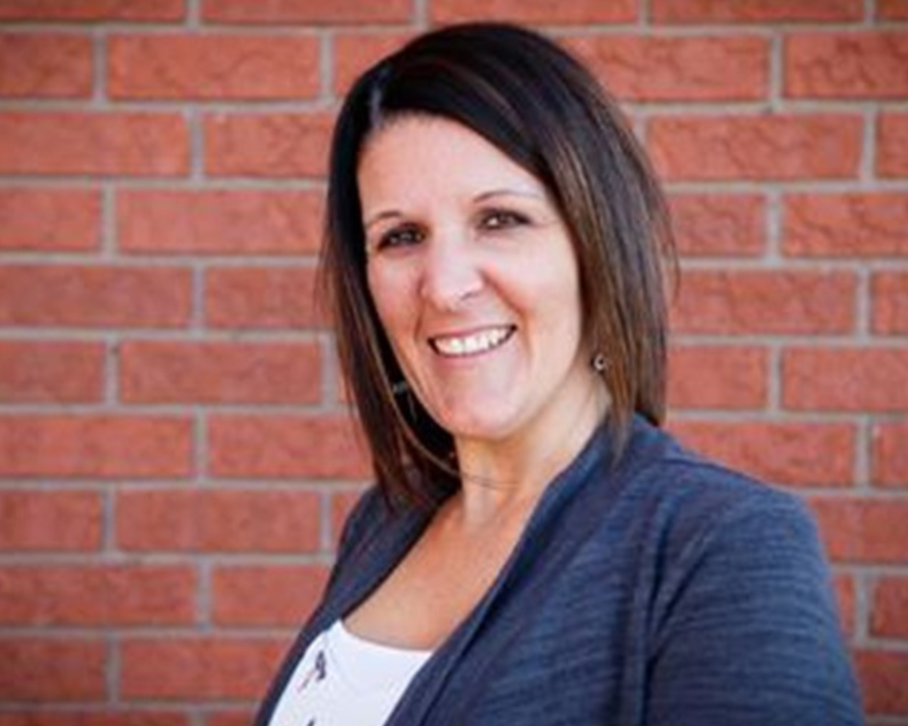 Smiling woman with shoulder-length dark hair, wearing a dark grey cardigan over a white top, standing in front of a red brick wall.