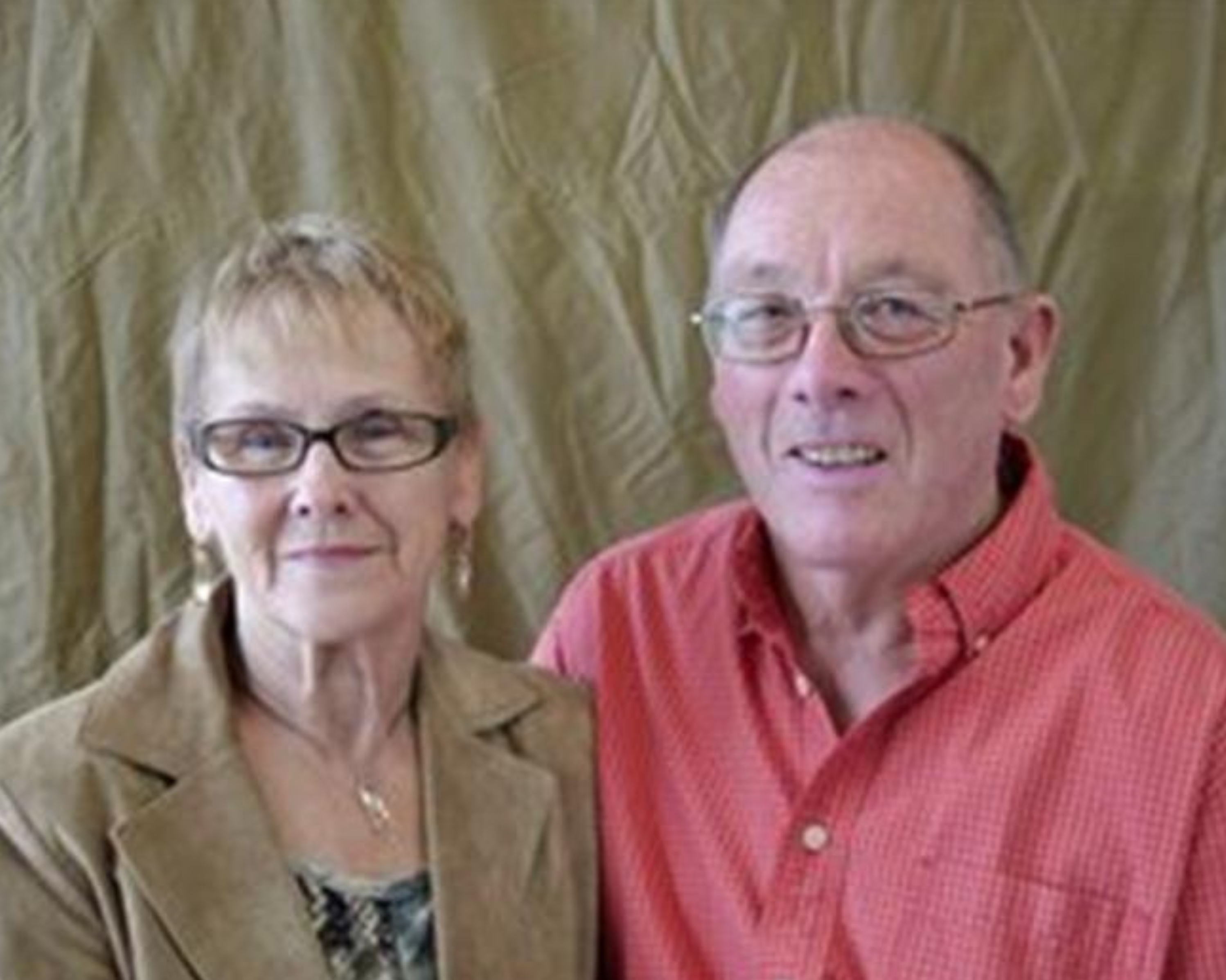 An older couple stands together, smiling at the camera. The woman on the left has short blonde hair, wears glasses, and is dressed in a beige blazer over a patterned blouse. The man on the right, also wearing glasses, is dressed in a red checkered shirt. The background is a plain beige curtain, creating a simple and warm setting for the portrait.
