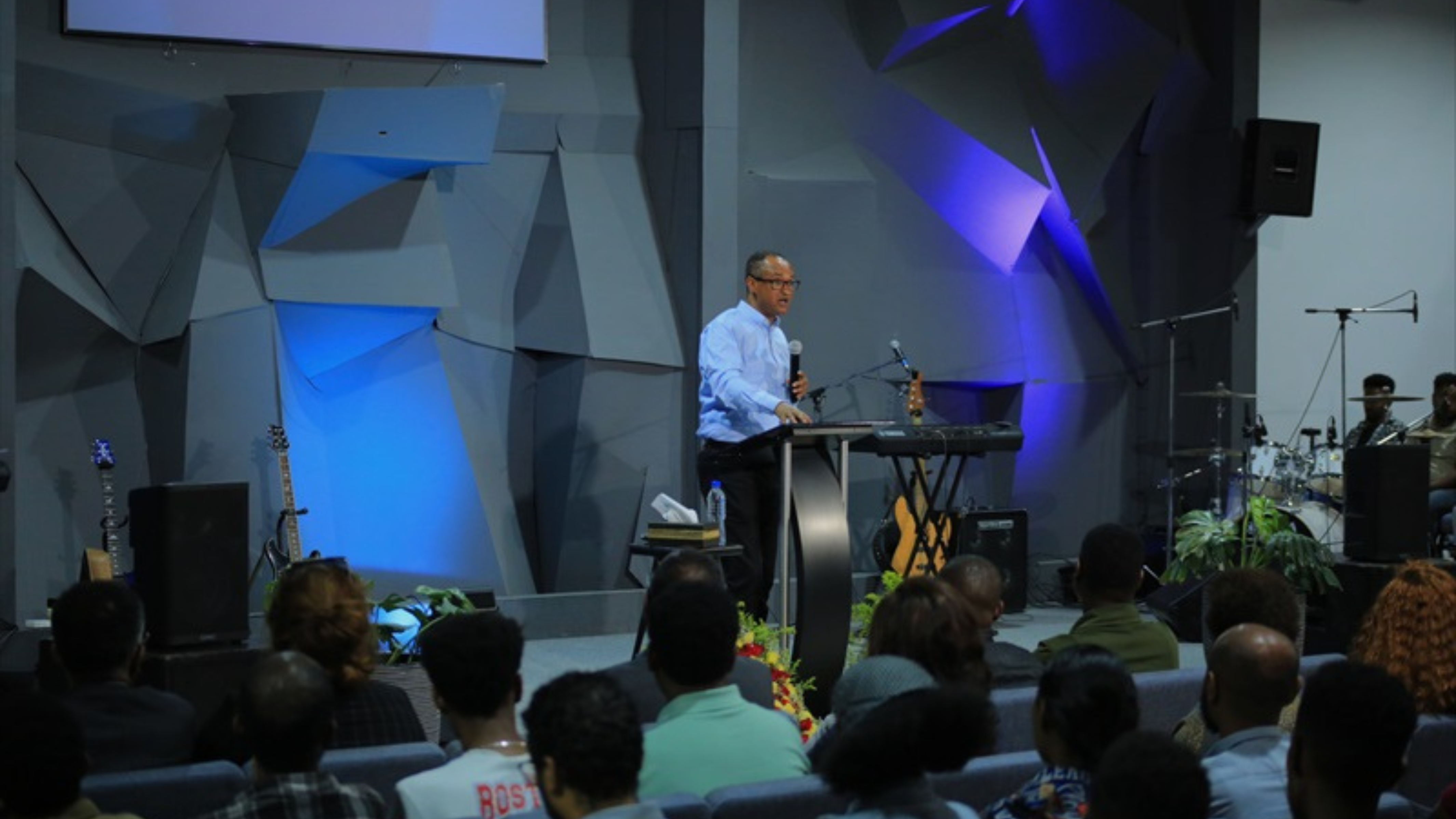 The image shows a speaker standing behind a lectern, addressing an audience in a modern church setting. The speaker is wearing a light blue shirt and is speaking into a microphone. The stage has a unique, angular backdrop with blue and purple lighting, creating a dynamic and contemporary atmosphere. The audience is seated in rows, attentively listening to the speaker. Musical instruments, including a guitar and drums, are visible on the stage, indicating a blend of preaching and worship in the service.