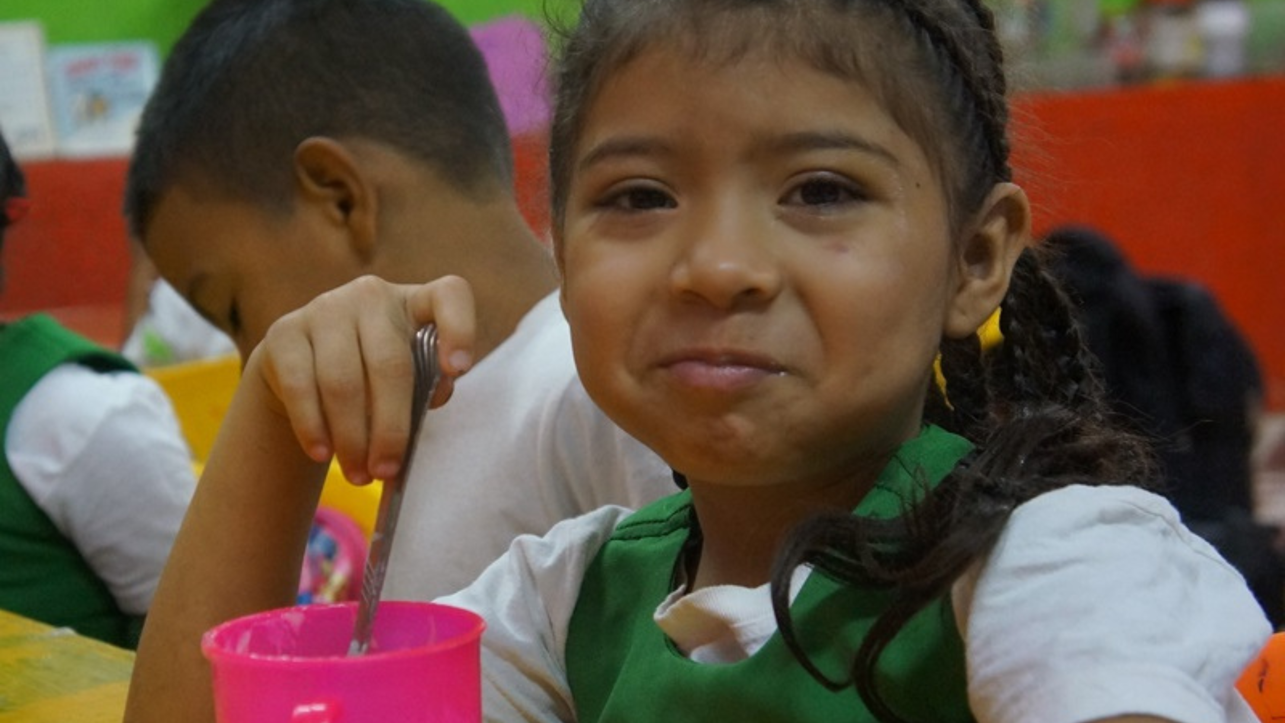 The image shows a young girl sitting at a table, smiling slightly as she enjoys a meal. She is holding a spoon and has a bright pink cup in front of her. She is wearing a white shirt and a green vest. Other children can be seen in the background, also engaged in their meals. The setting appears to be a cheerful, colorful room, likely a classroom or a community center, with a green and red painted wall. The girl's expression is warm and content, giving a sense of comfort and care.