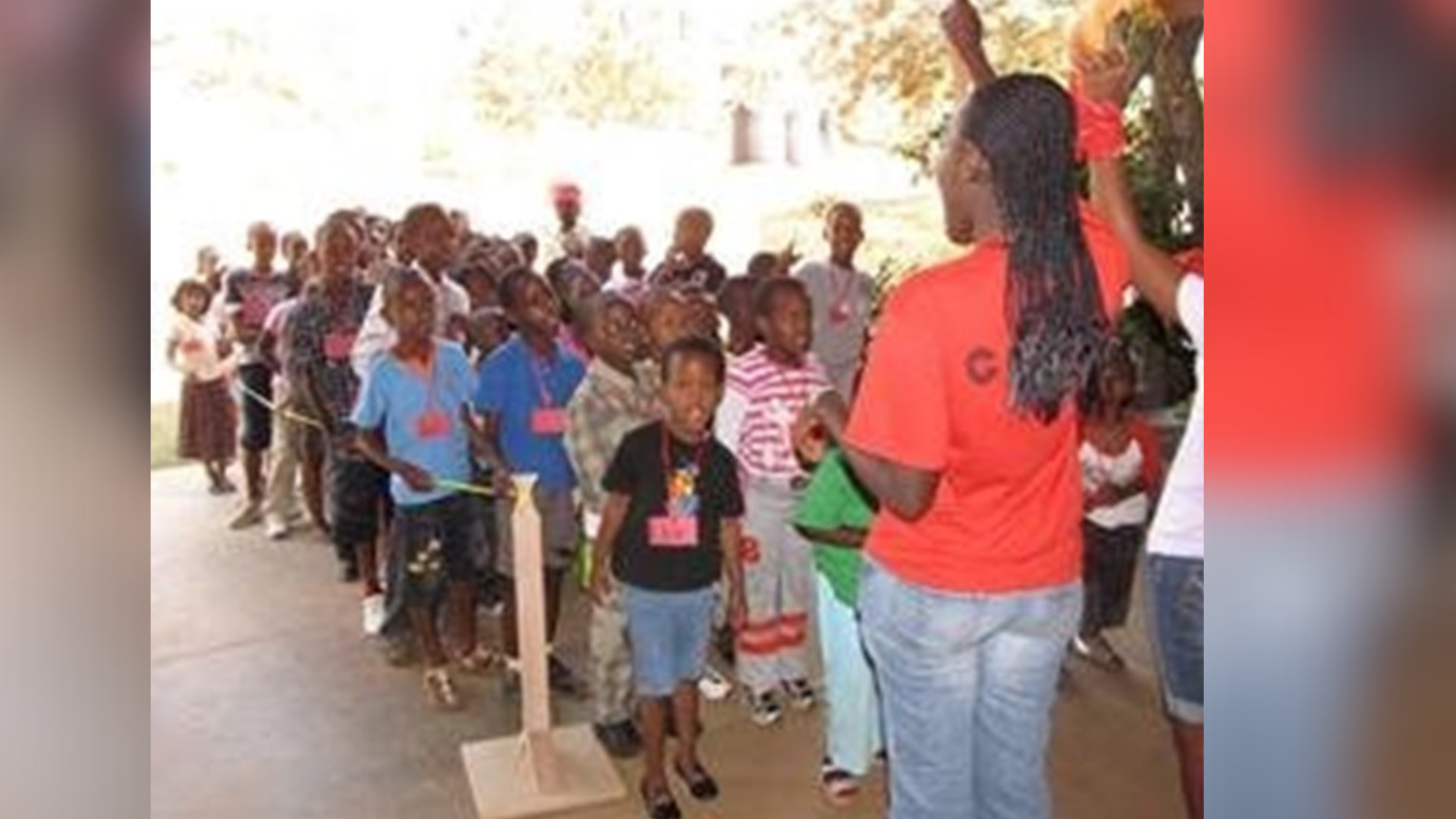 A lively group of children and a few adults in an outdoor setting, participating in a group activity. The children, wearing colorful casual clothes and name tags, are actively engaging and looking towards the adult leaders who are coordinating the event. The leaders, in bright orange shirts, are encouraging the children with raised hands and enthusiastic expressions.