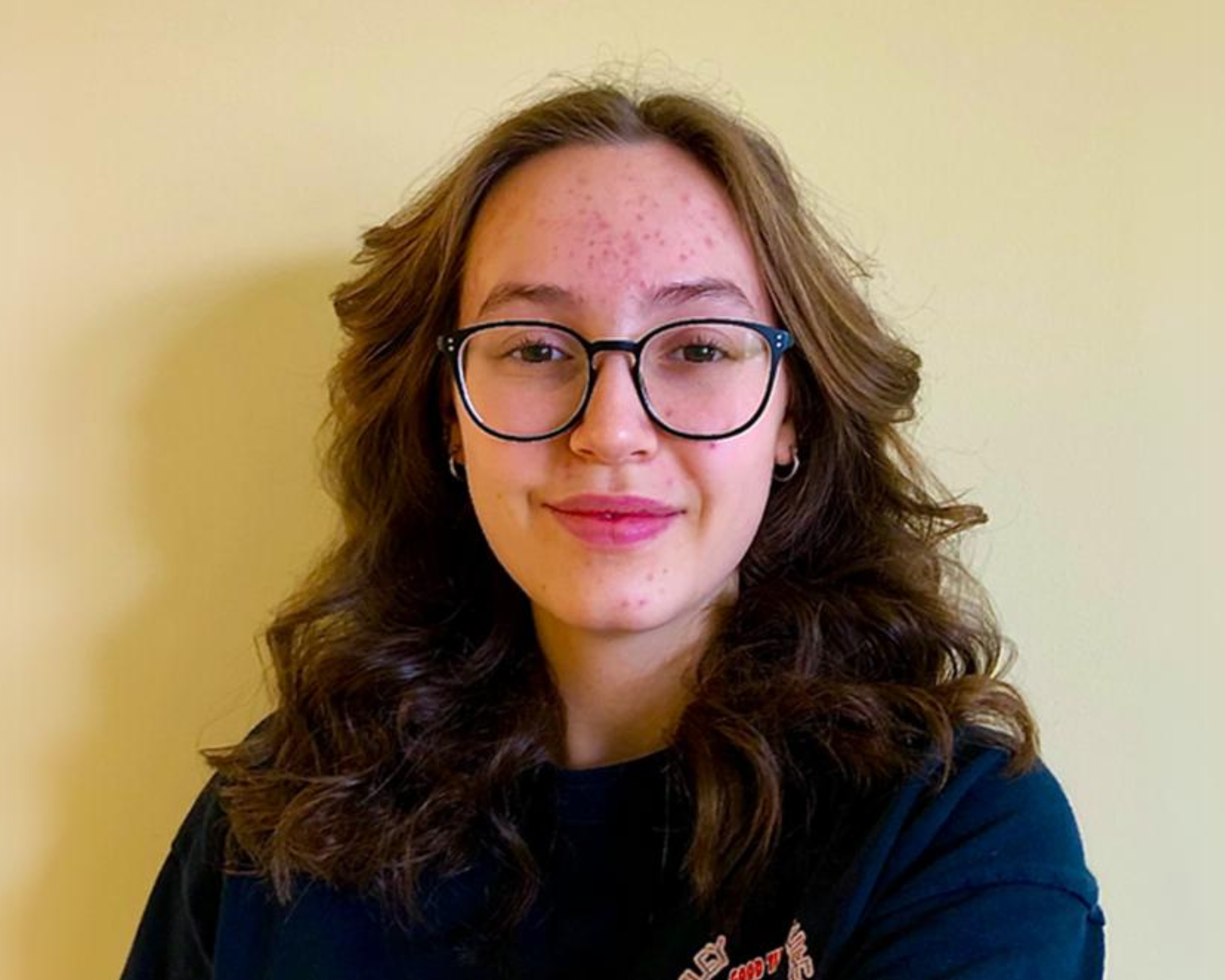 A young woman with wavy brown hair is standing against a plain yellow background. She is wearing glasses and a black shirt, and has a gentle, confident smile. Her hair frames her face, and she has a natural, relaxed expression. The image is simple and well-lit, focusing on her face.