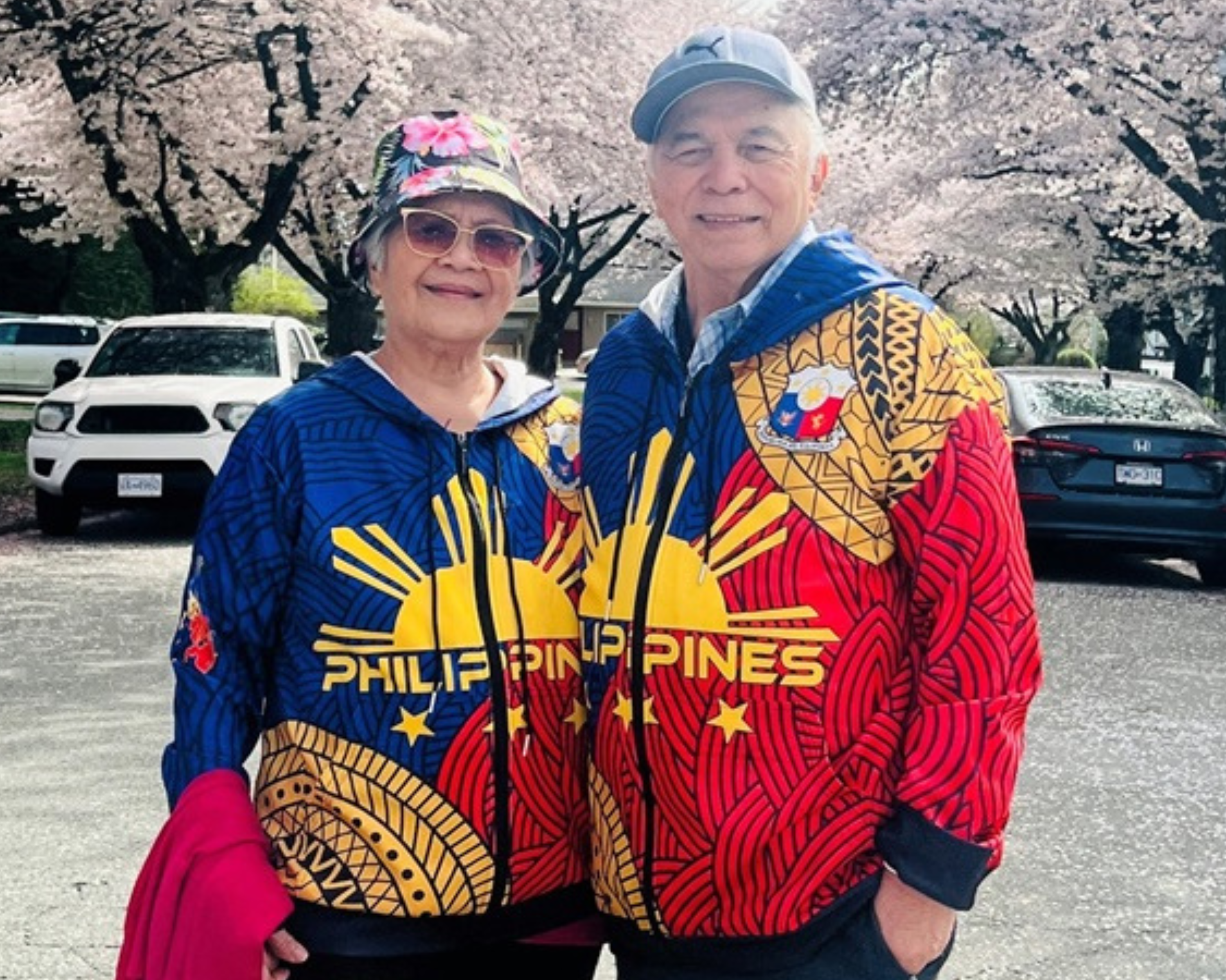 An older couple stands together on a street lined with blooming cherry blossom trees. They are both wearing matching colorful jackets with the word "Philippines" and the sun and stars from the Philippine flag prominently displayed. The woman is also wearing a floral hat and sunglasses, while the man is wearing a cap. They are smiling and appear to be enjoying a beautiful day outdoors, with parked cars and houses visible in the background.