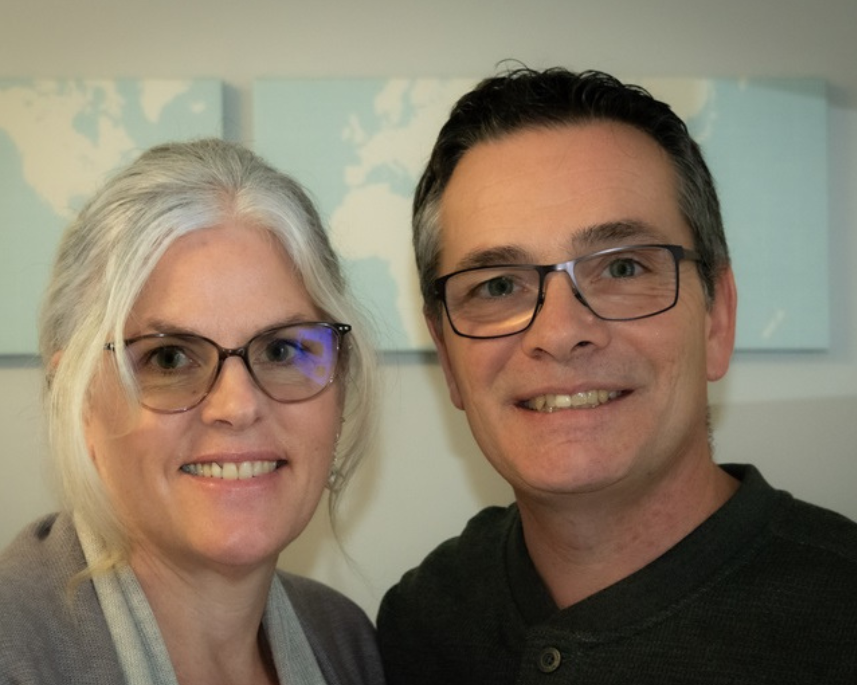 A couple smiling indoors with a map of the world as a background. The woman on the left has grey hair and wears glasses, and a light-coloured cardigan. The man on the right, also wearing glasses, has short dark hair with some grey and is dressed in a dark green sweater. Both appear content and happy.