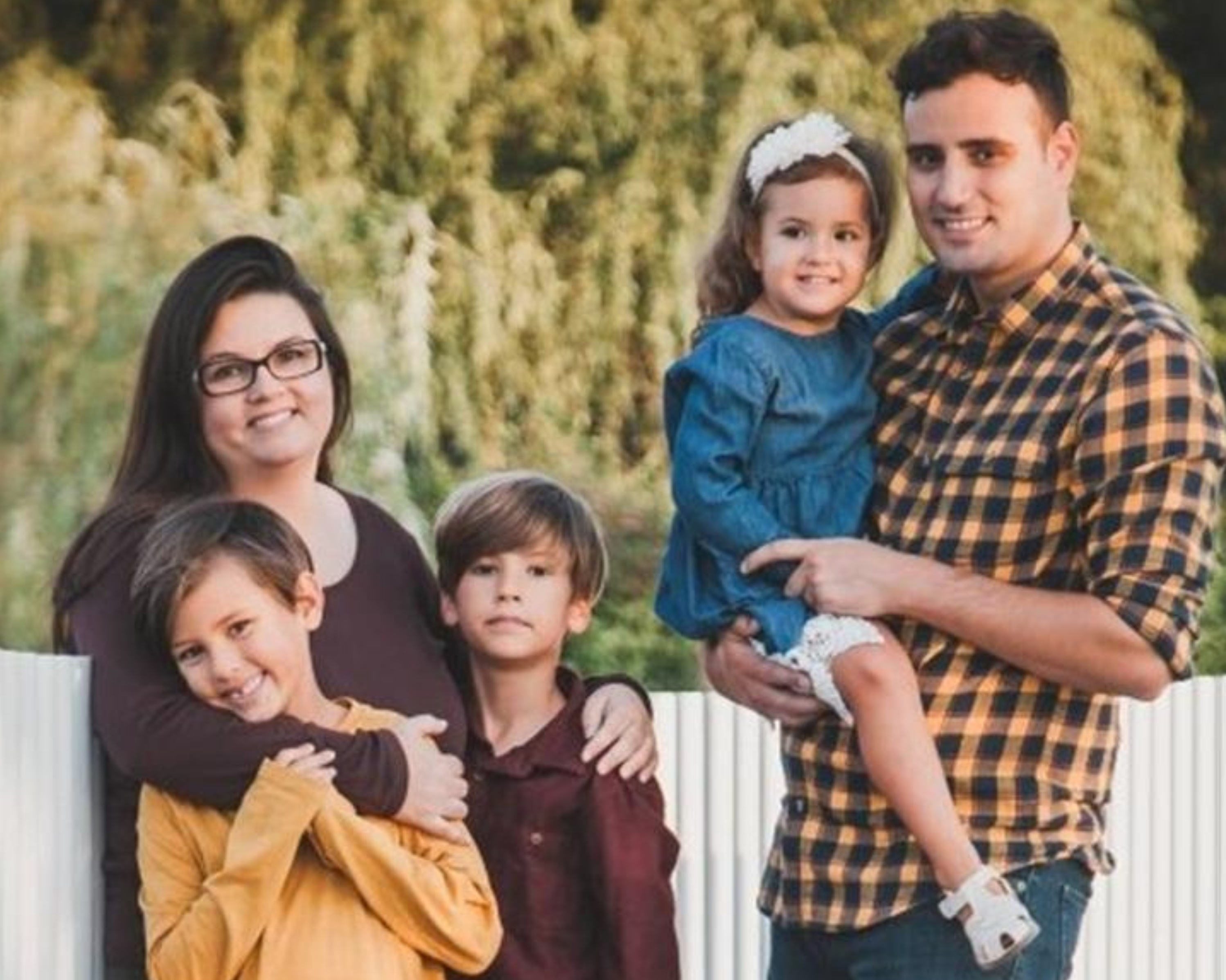 A family of five stands together outdoors in front of a white fence with greenery in the background. The mother on the left is wearing glasses and a dark sweater, holding a young boy in a mustard-coloured shirt. Next to them stands another young boy in a maroon shirt. The father on the right is wearing a yellow and black checkered shirt, holding a little girl in a blue dress with a white headband. The family looks happy and relaxed, enjoying the time together.