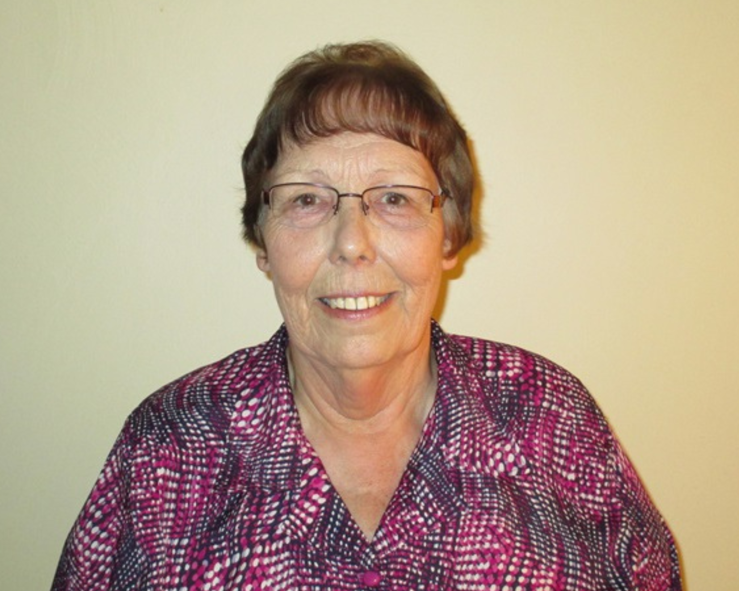 An elderly woman is smiling at the camera while standing against a plain light-coloured wall. She is wearing glasses and a patterned blouse in shades of pink, black, and white. Her short brown hair is neatly styled, and she exudes a warm, approachable demeanour.