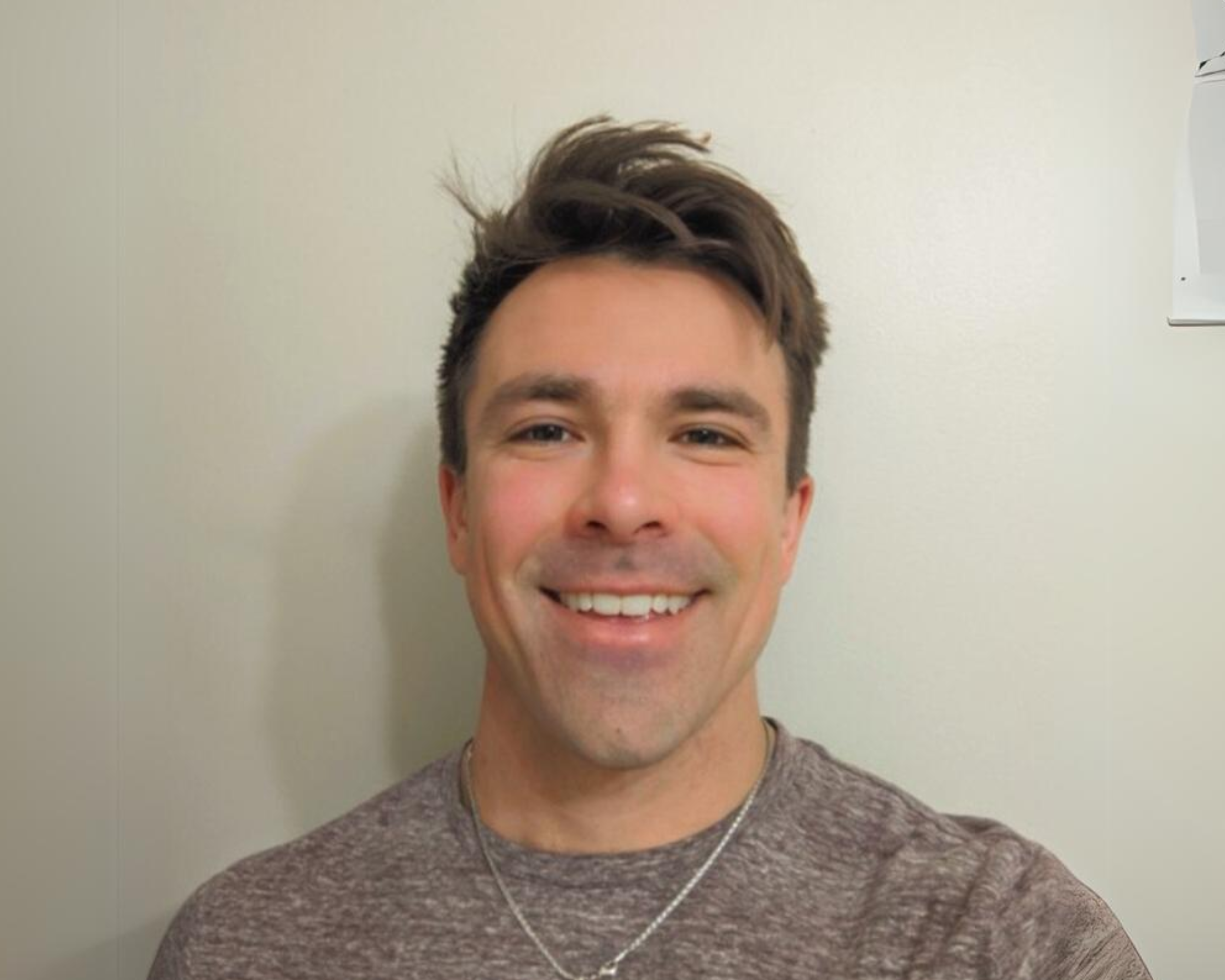 A smiling man with short brown hair is standing against a plain light-coloured background. He is wearing a maroon t-shirt and a silver cross necklace. His expression is friendly and approachable, and the photo focuses on his face and upper body.