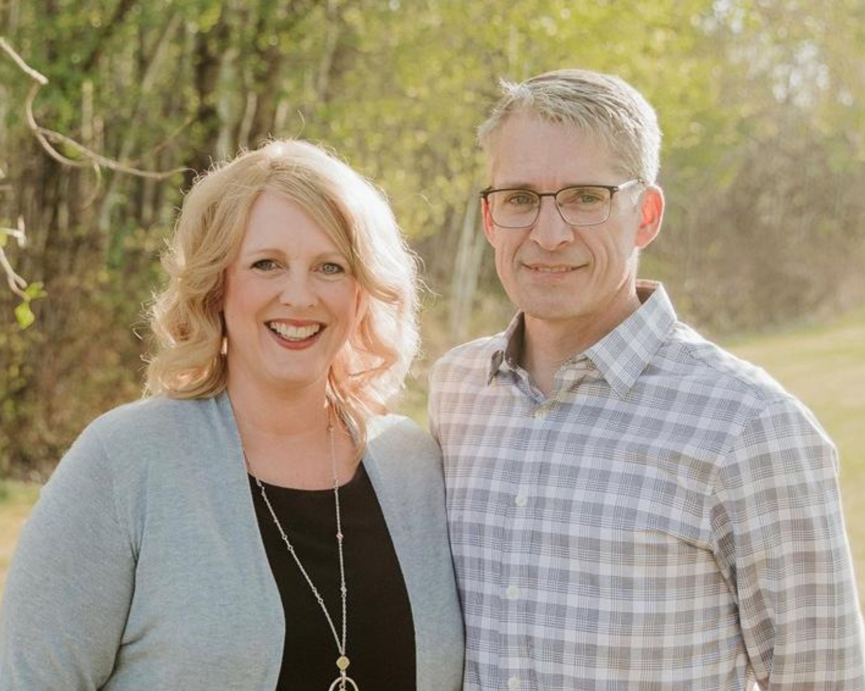 A man and woman stand side by side, smiling warmly at the camera. They are outdoors, with a background of trees and greenery softly lit by natural sunlight. The woman wears a light grey cardigan and a necklace, while the man is dressed in a checkered shirt with glasses.