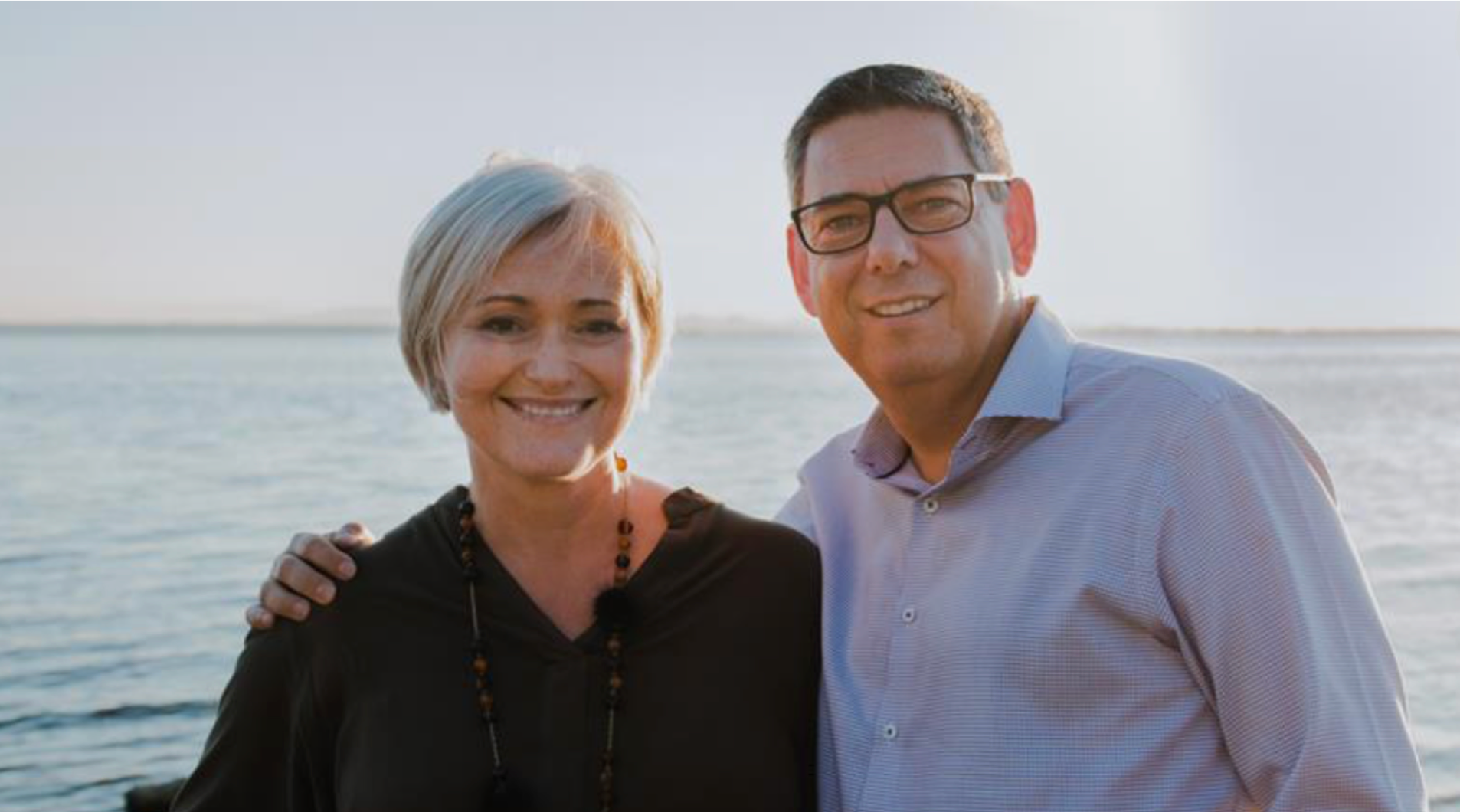 photo of couple smiling at camera behind view of a lake