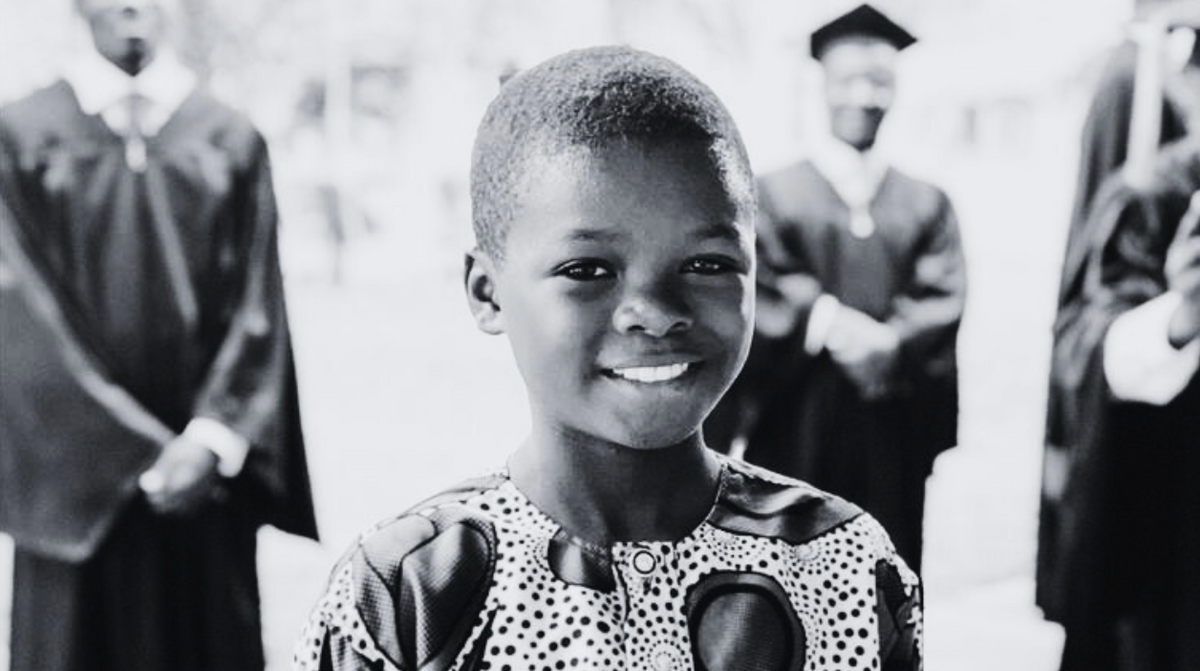 black and white photo of young child smiling at camera behind is a blurred photo of a few graduates in their ceremonial gowns and hats.