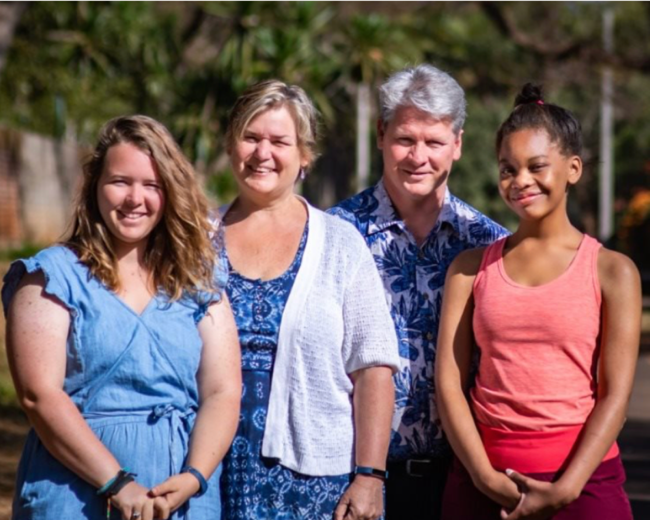 photo of couple and two young adult girls smiling in front of camera behind trees and a street view.