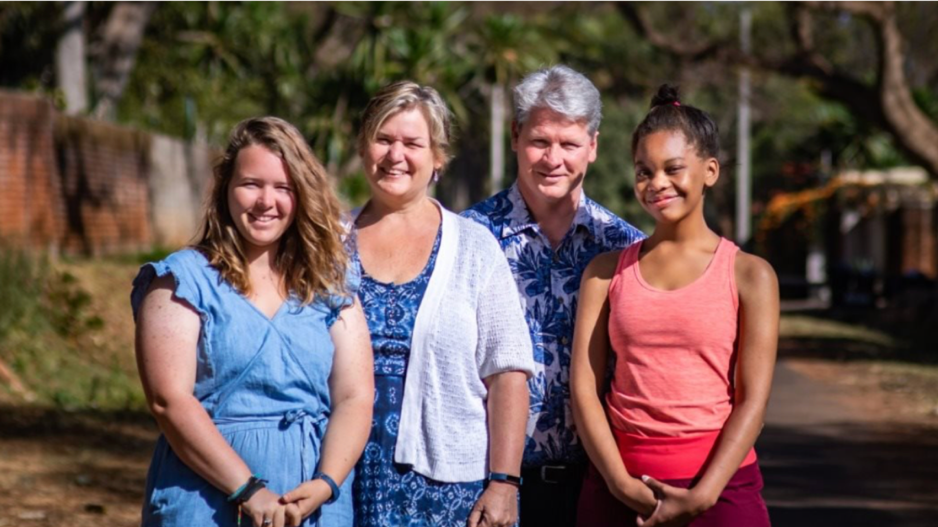 photo of couple and two young adult girls smiling in front of camera behind trees and a street view.