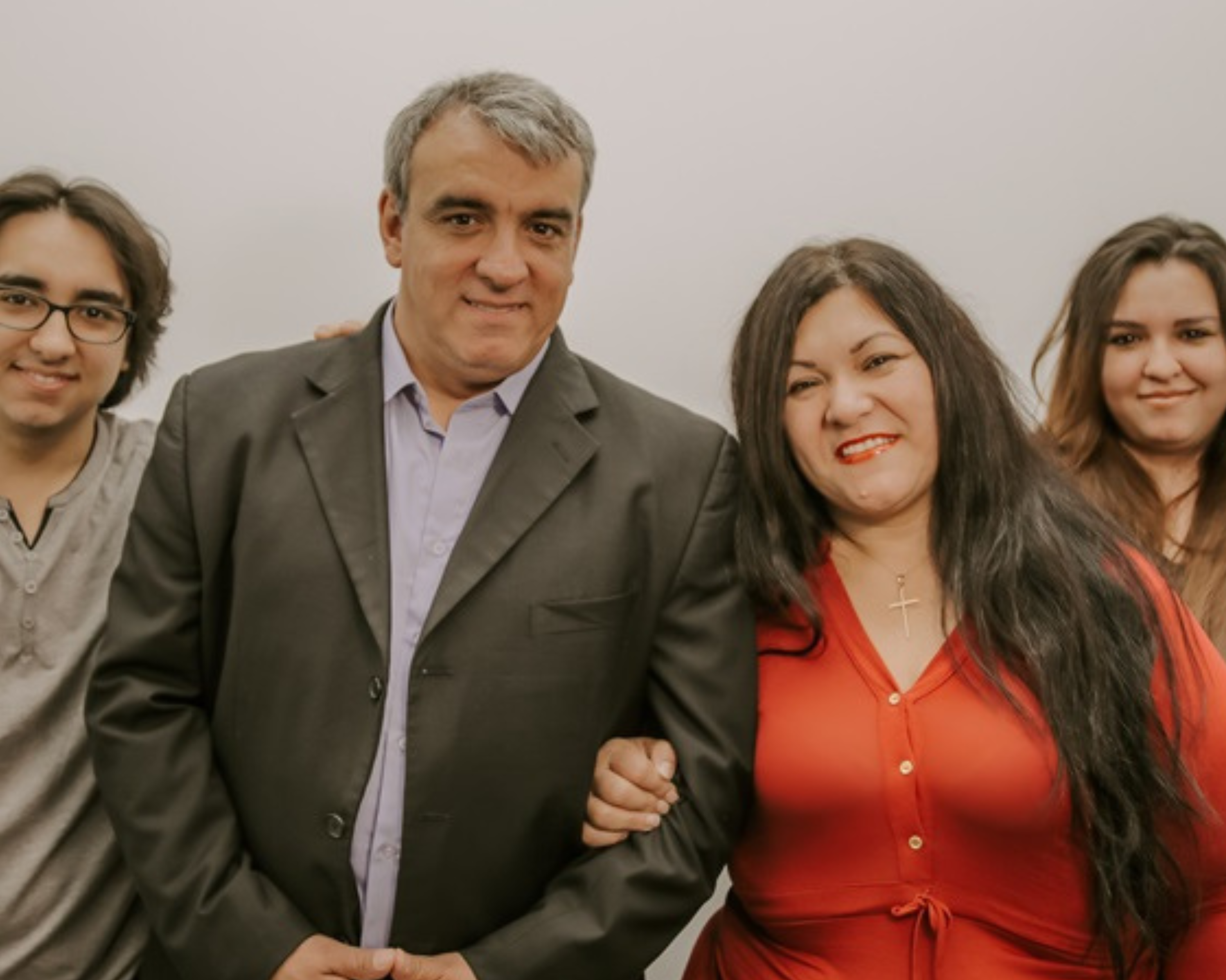 Portrait of a family consisting of four members standing closely together against a neutral background. From left to right: a young man with shoulder-length hair wearing a gray shirt, a middle-aged man in a dark suit and blue shirt, a woman in a bright red blouse, and a young woman in a black jacket. All are smiling warmly at the camera.