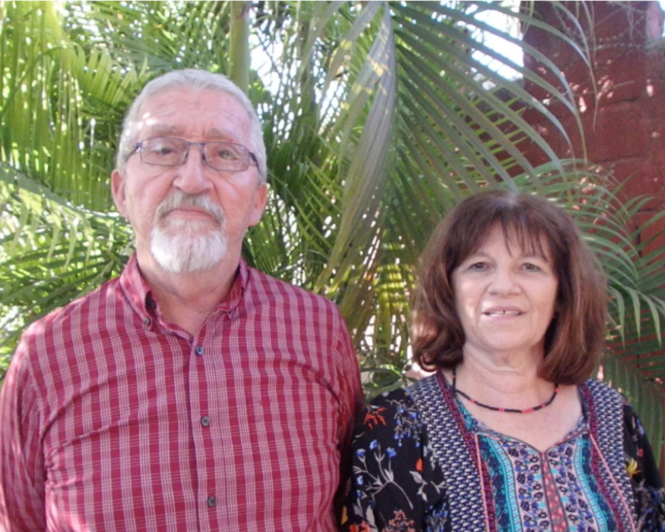Portrait of an elderly couple standing outdoors with tropical foliage in the background. The man on the left wears glasses and a red checked shirt, and the woman on the right has shoulder-length brown hair and is dressed in a dark floral blouse. Both are smiling slightly and looking directly at the camera, with a rustic, colourful wall partially visible behind them.