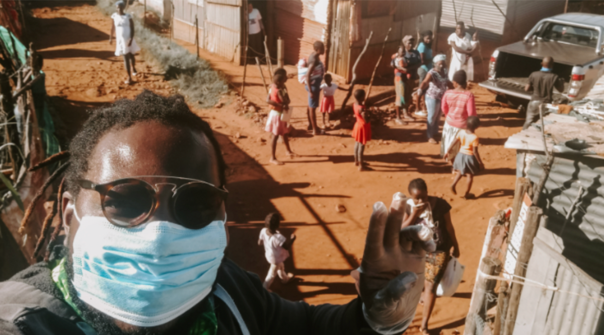 photo of GW in a mask showing peace sign at camera behind is a village with people walking on the street.