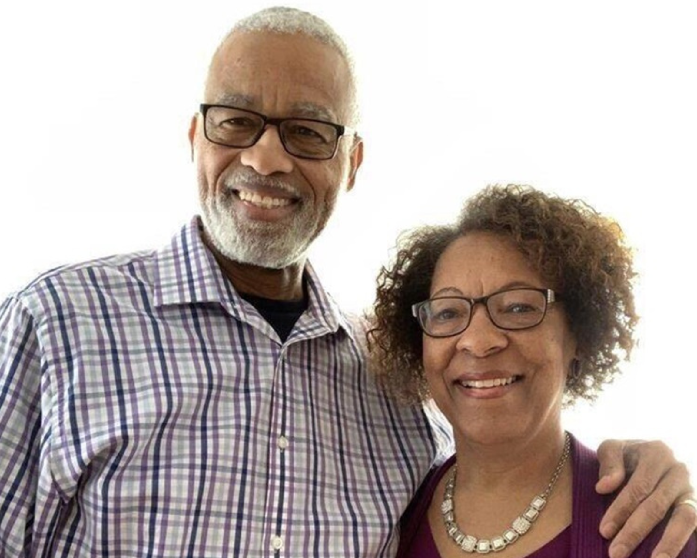 Portrait of an elderly couple smiling at the camera. The man, wearing glasses and a plaid shirt, stands slightly behind the woman who is dressed in a purple cardigan over a maroon top. Both exhibit warm, welcoming expressions, with a plain, light background enhancing their figures.