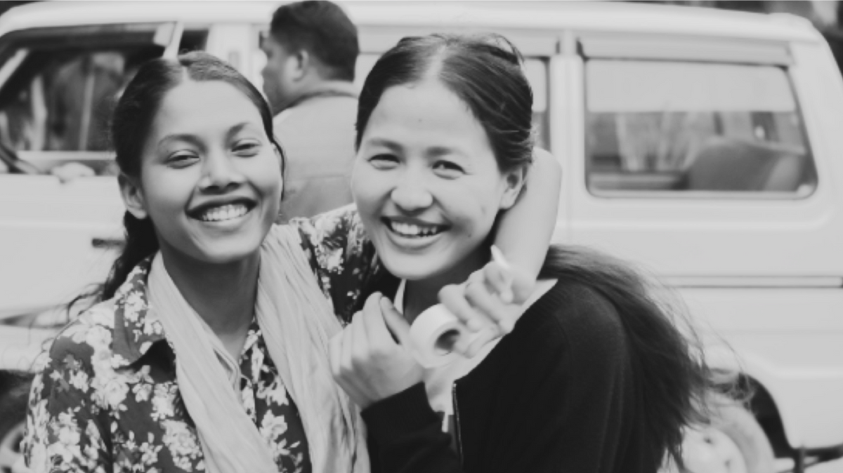 black and white photo of two women laughing, and smiling at camera behind a white vehicle.
