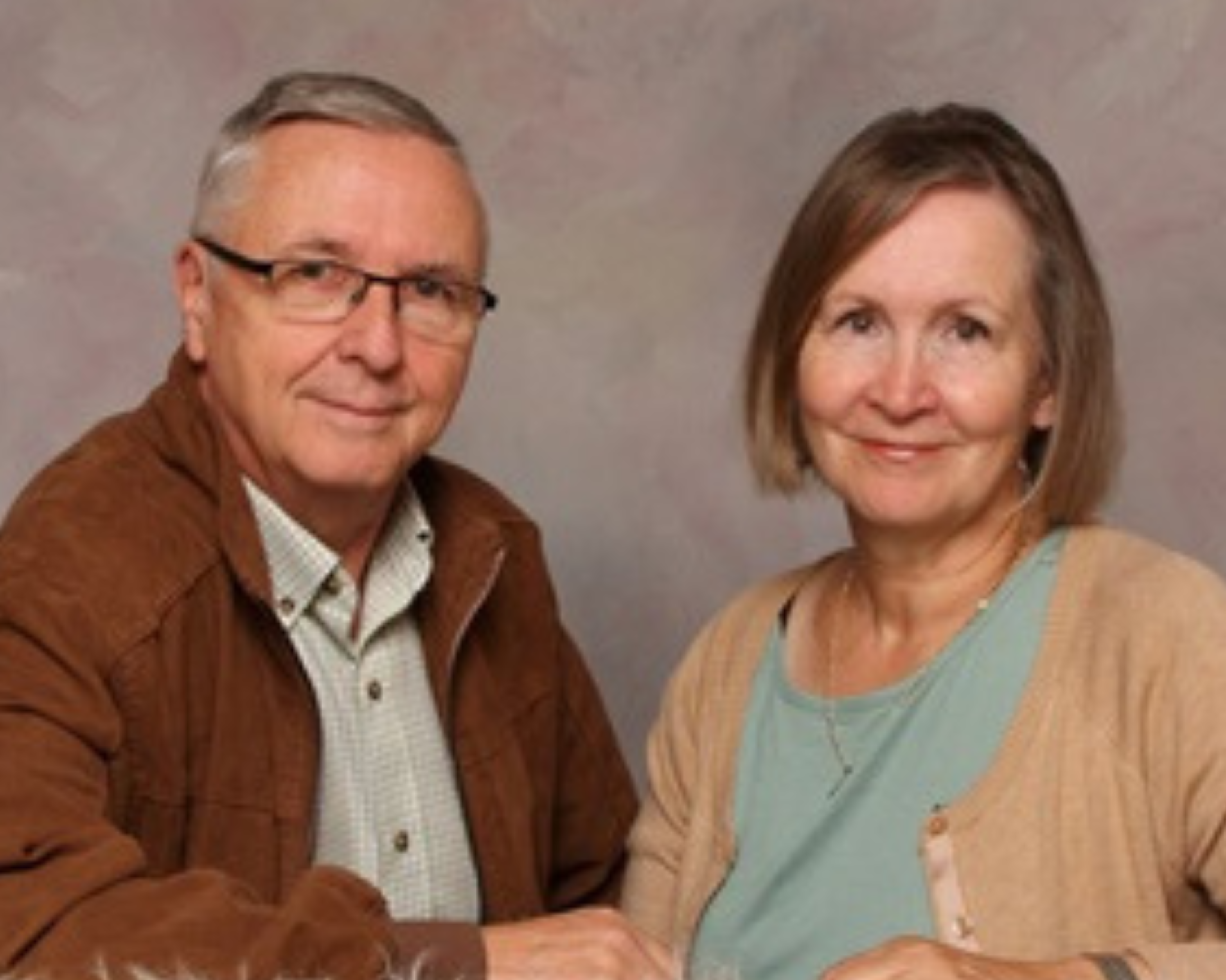 Portrait of an elderly couple seated close together, looking at the camera with a calm expression. The man wears glasses, a brown jacket over a plaid shirt, and the woman is dressed in a beige cardigan over a light blue top. They are seated against a soft, mottled tan background, resting their arms on a fluffy white surface.