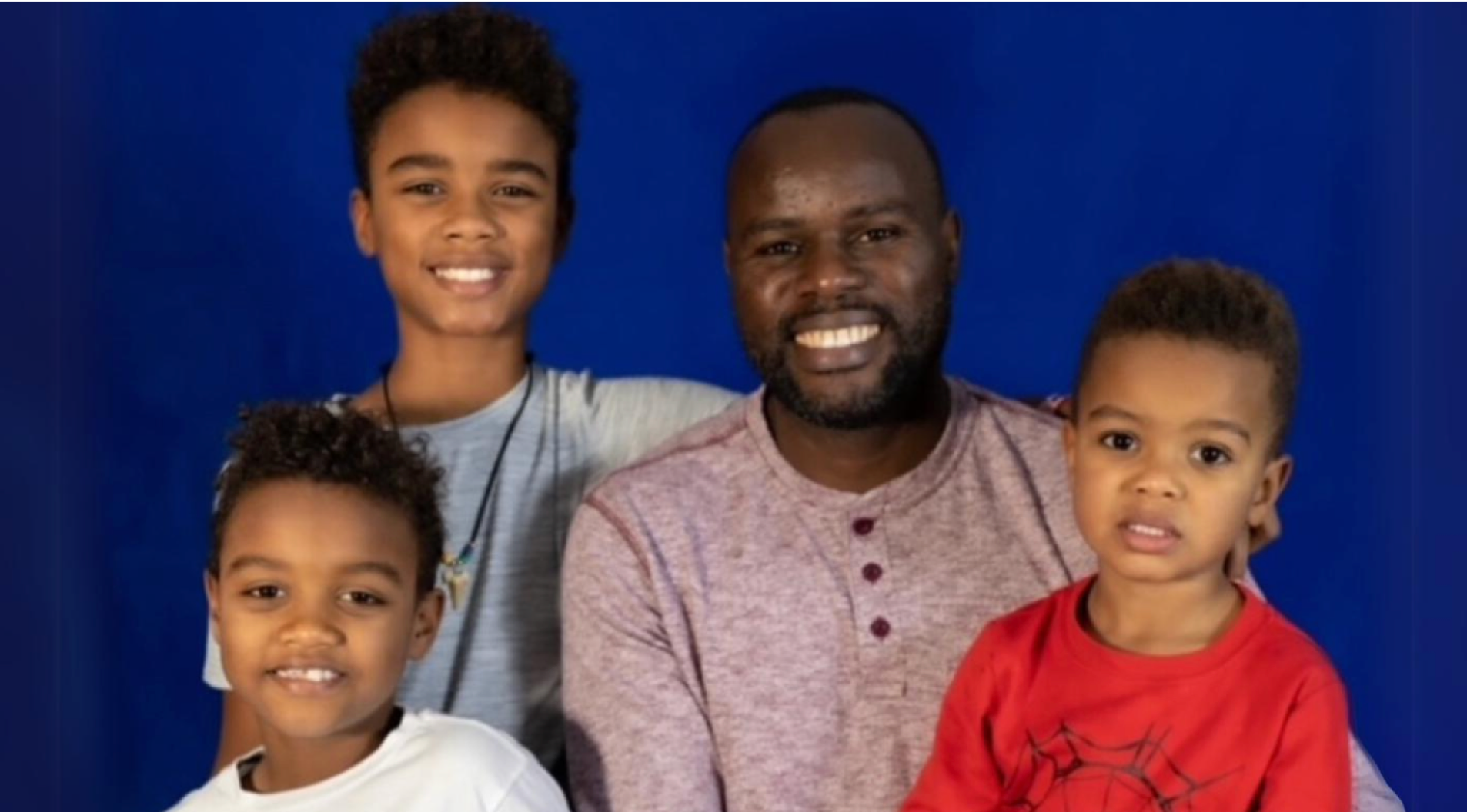 photo of man and three young boys smiling at the camera behind blue wall