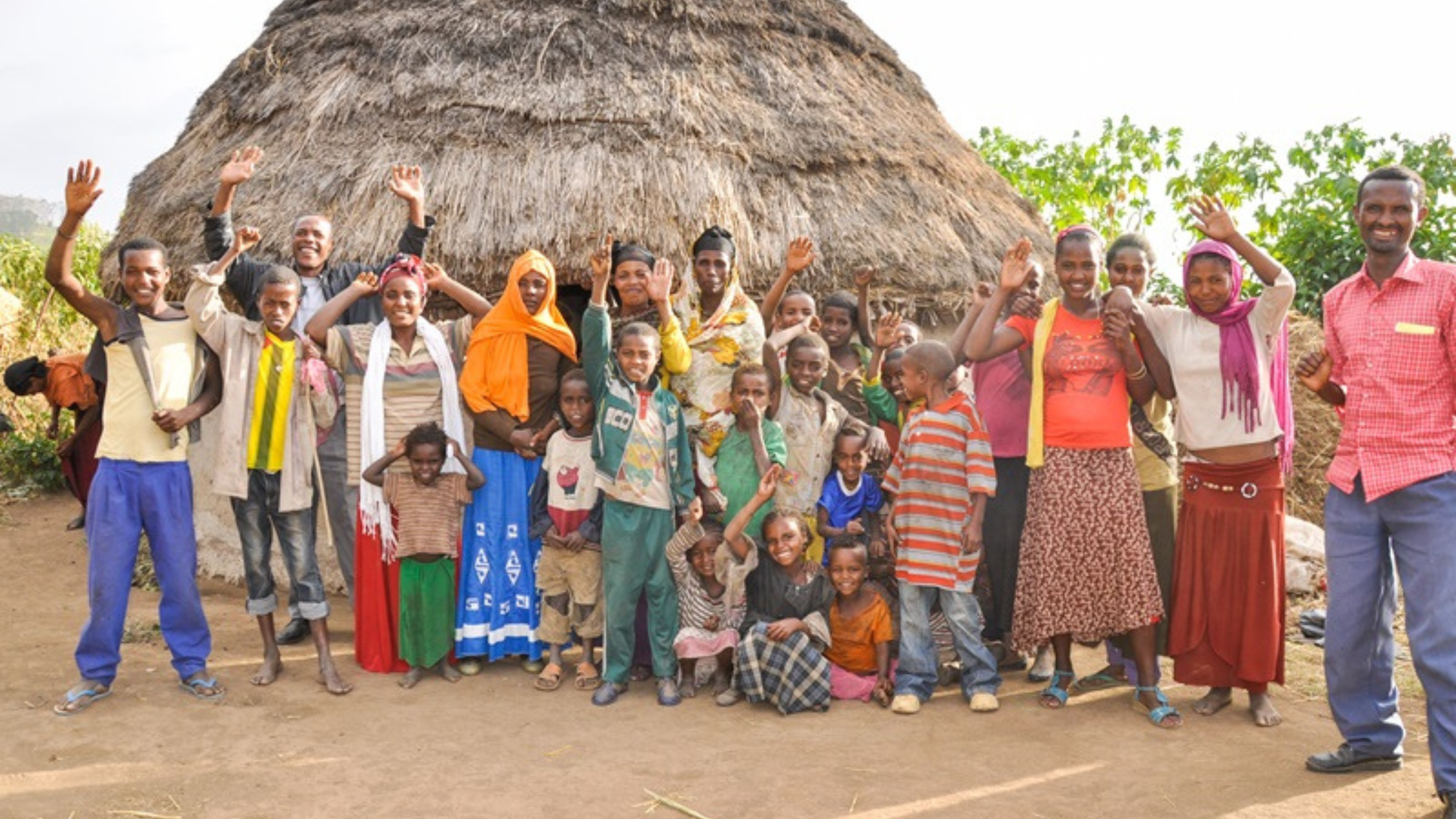 The image shows a large group of men, women, and children smiling and waving at the camera, standing in front of a traditional round, thatched-roof hut. The people are wearing a variety of colorful clothing, and the scene suggests a rural village setting. The group appears joyful and welcoming, with children in the front and adults standing behind them. The background features greenery, giving the image a warm and communal atmosphere, reflecting unity and a strong sense of community.