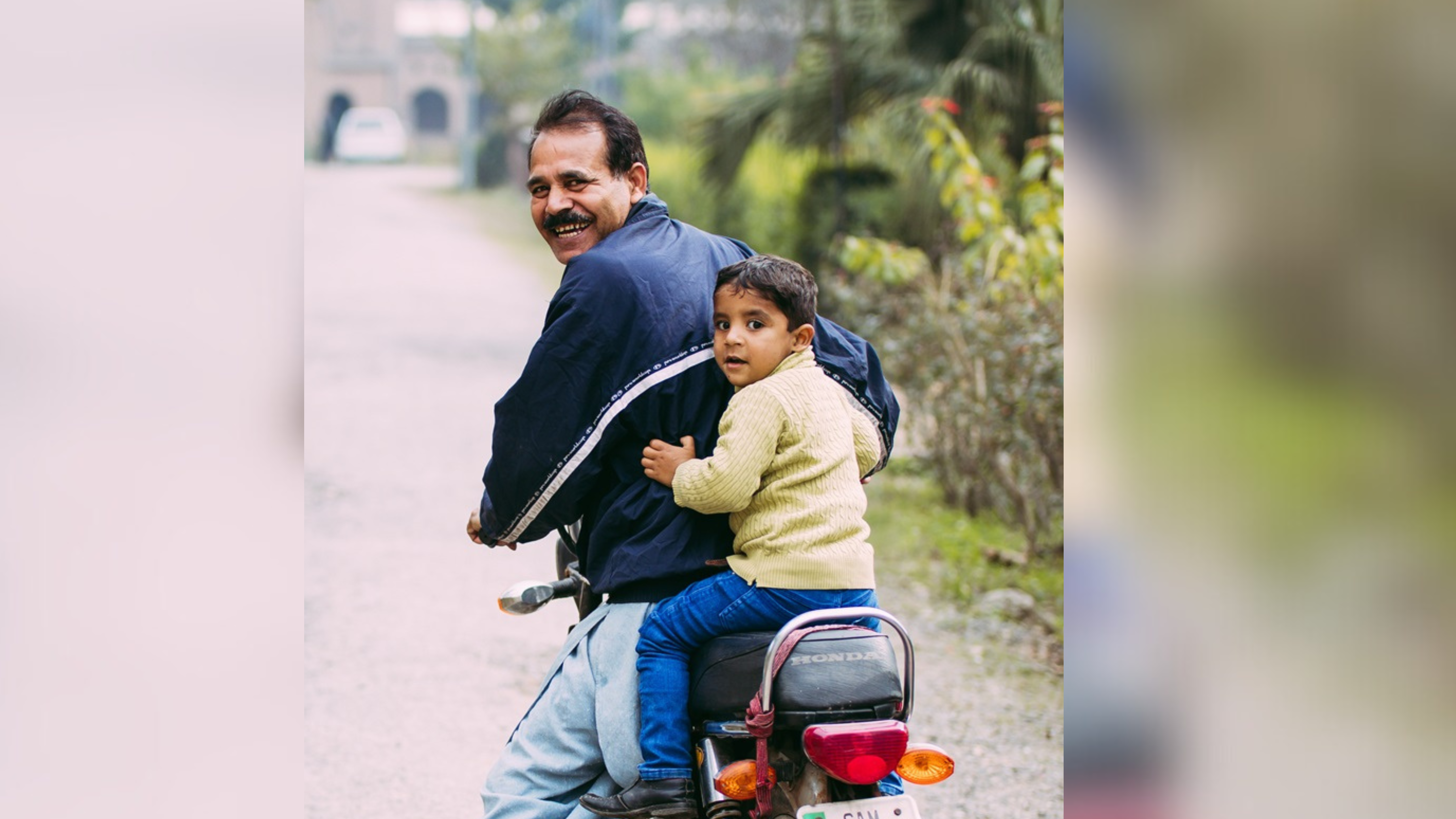 A cheerful man and a young boy are riding a motorcycle down a quiet, tree-lined road. The man, wearing a navy jacket and smiling over his shoulder, has the boy securely wrapped around him. The boy, in a light sweater, looks towards the camera with a cautious expression.