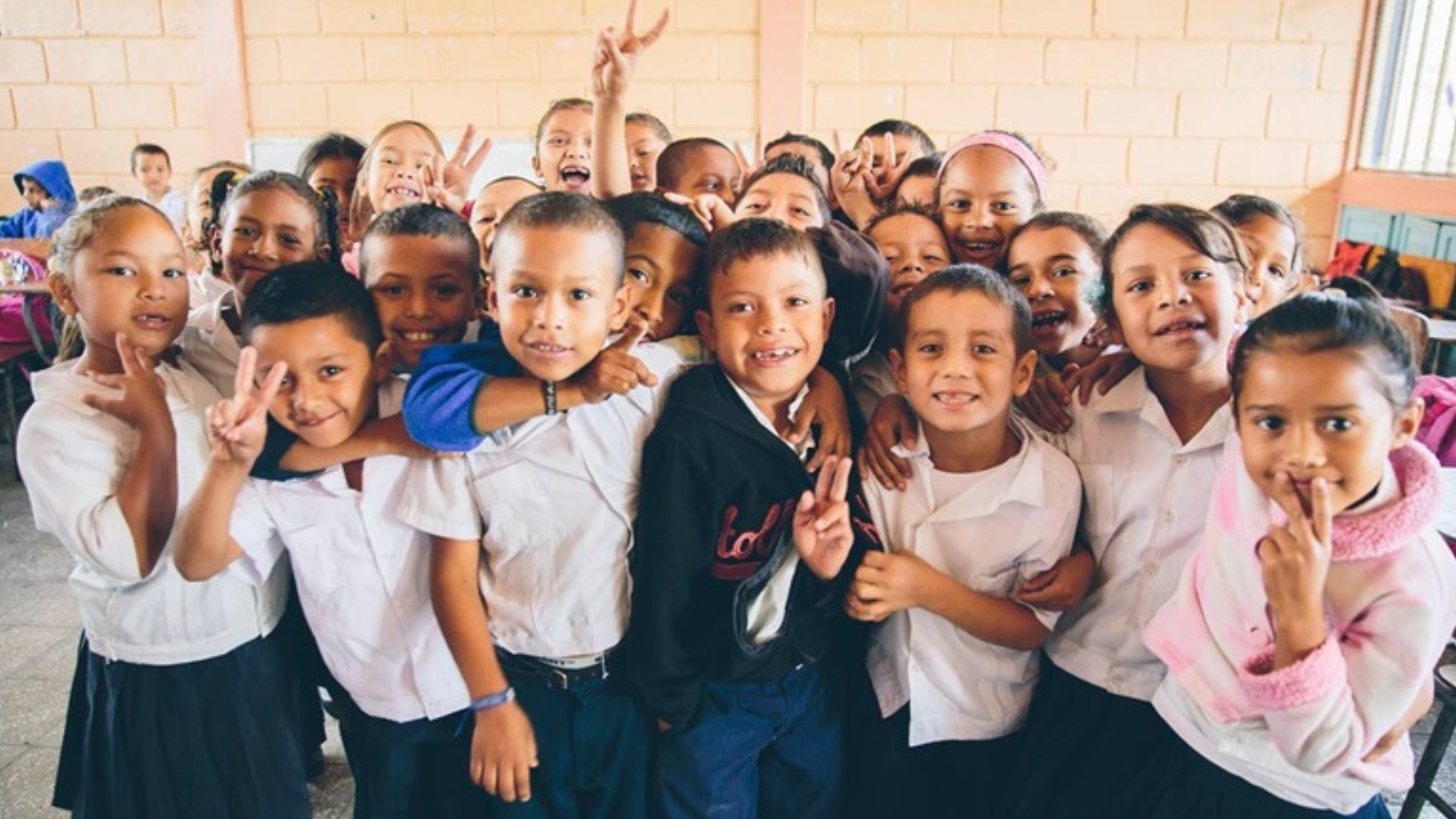 A lively group of elementary school children in a classroom setting, showing a mix of boys and girls dressed in school uniforms. The kids, displaying a range of joyful expressions and playful poses, are clustered together. Some are holding up peace signs, others are smiling broadly at the camera, and a few are waving. The background features classroom elements like desks and a chalkboard, adding to the lively educational environment.